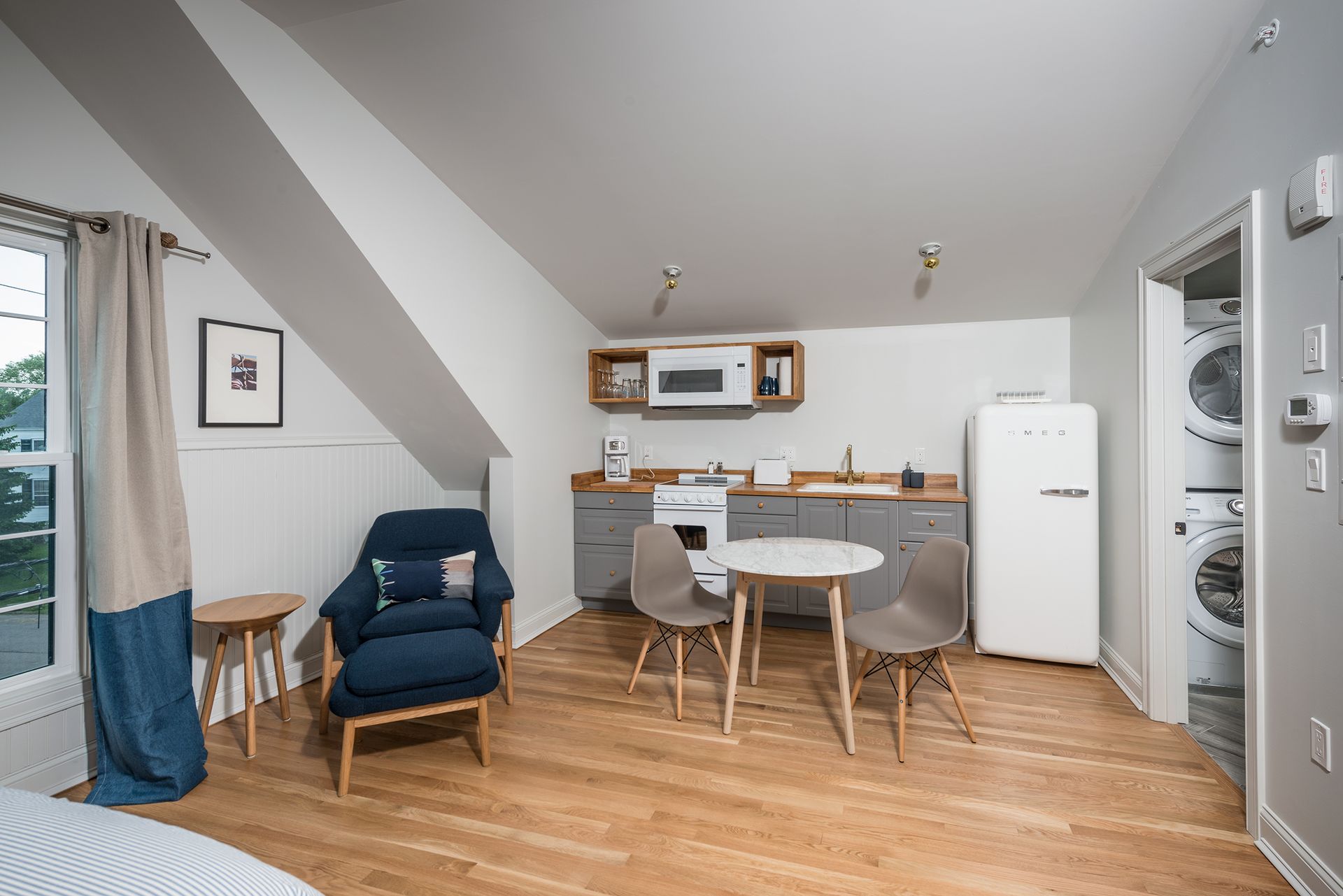 Small studio apartment with dining table, kitchenette, blue chair, bed, and stacked washer-dryer