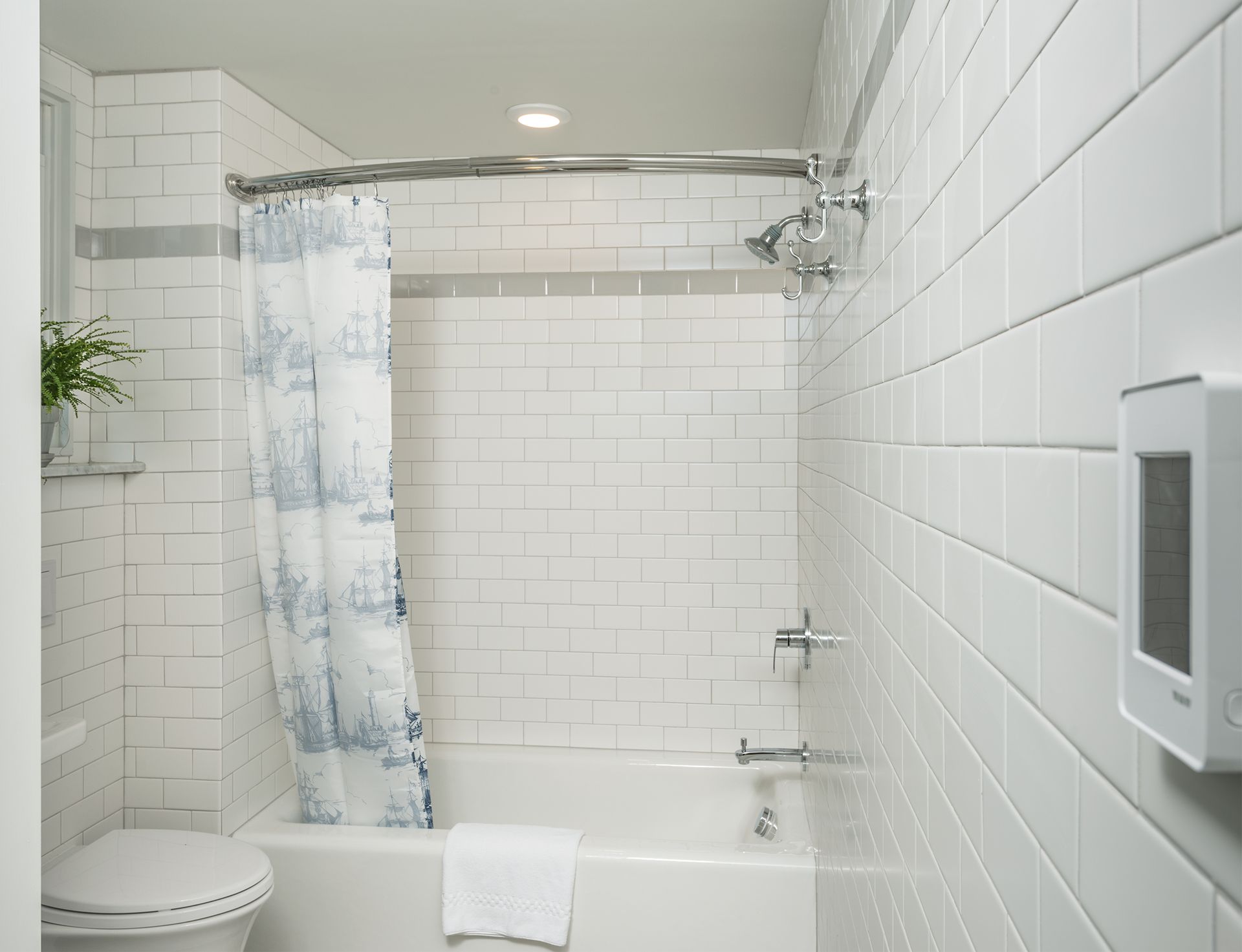 White tiled bathroom with bathtub, shower curtain, toilet, and wall-mounted soap dispenser