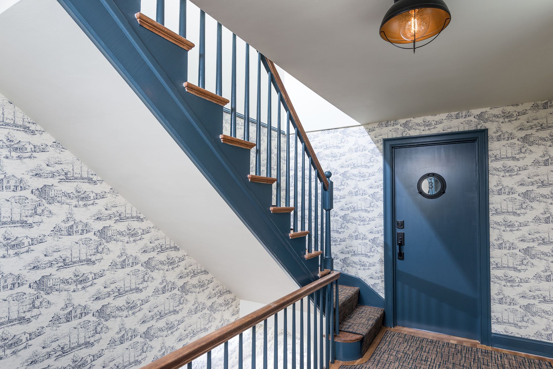 Blue stairwell with patterned wallpaper, wooden steps, and a blue door with a round window.
