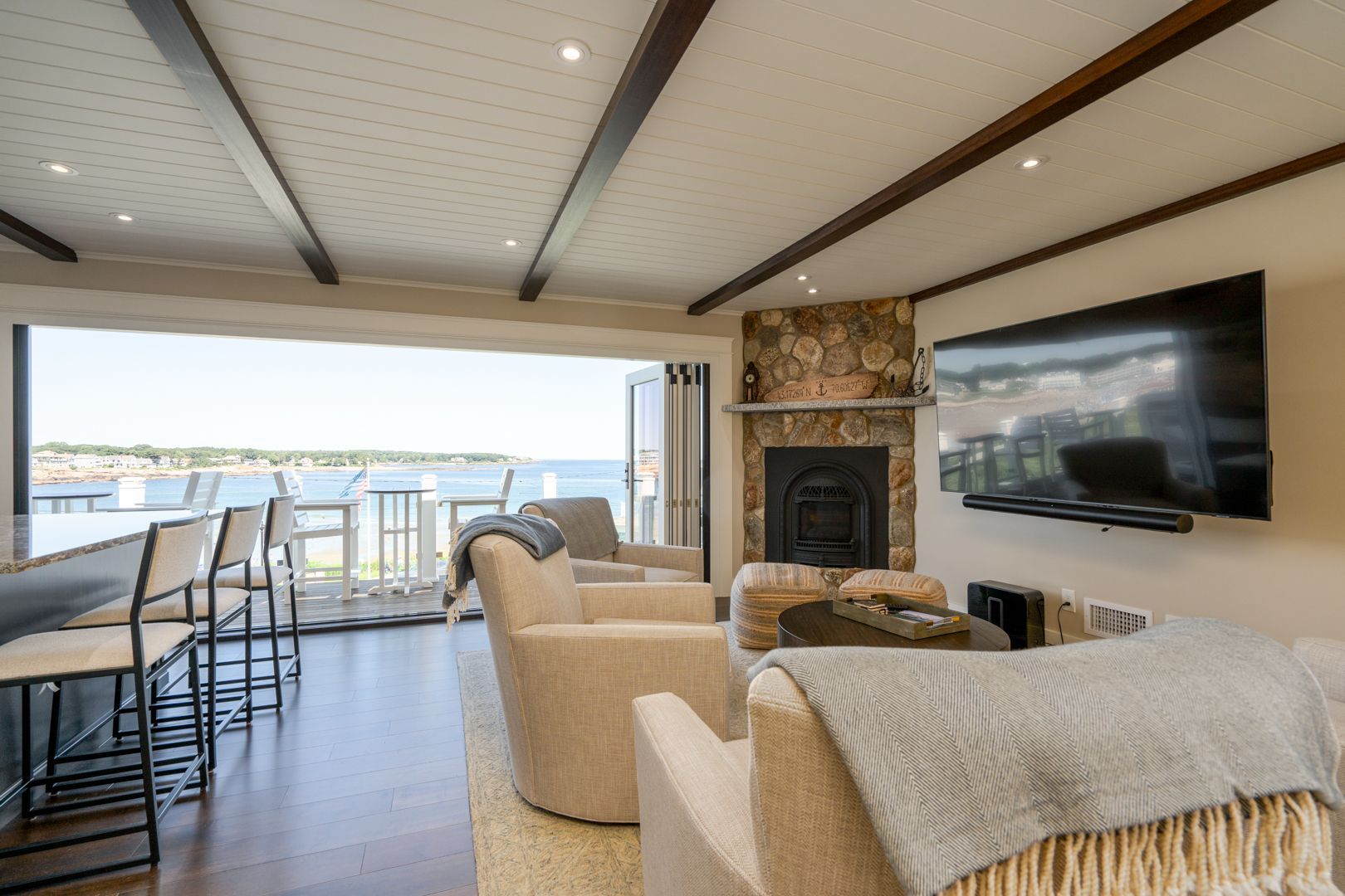 Bright living room with stone fireplace, wall TV, bar stools, and ocean view through large sliding doors