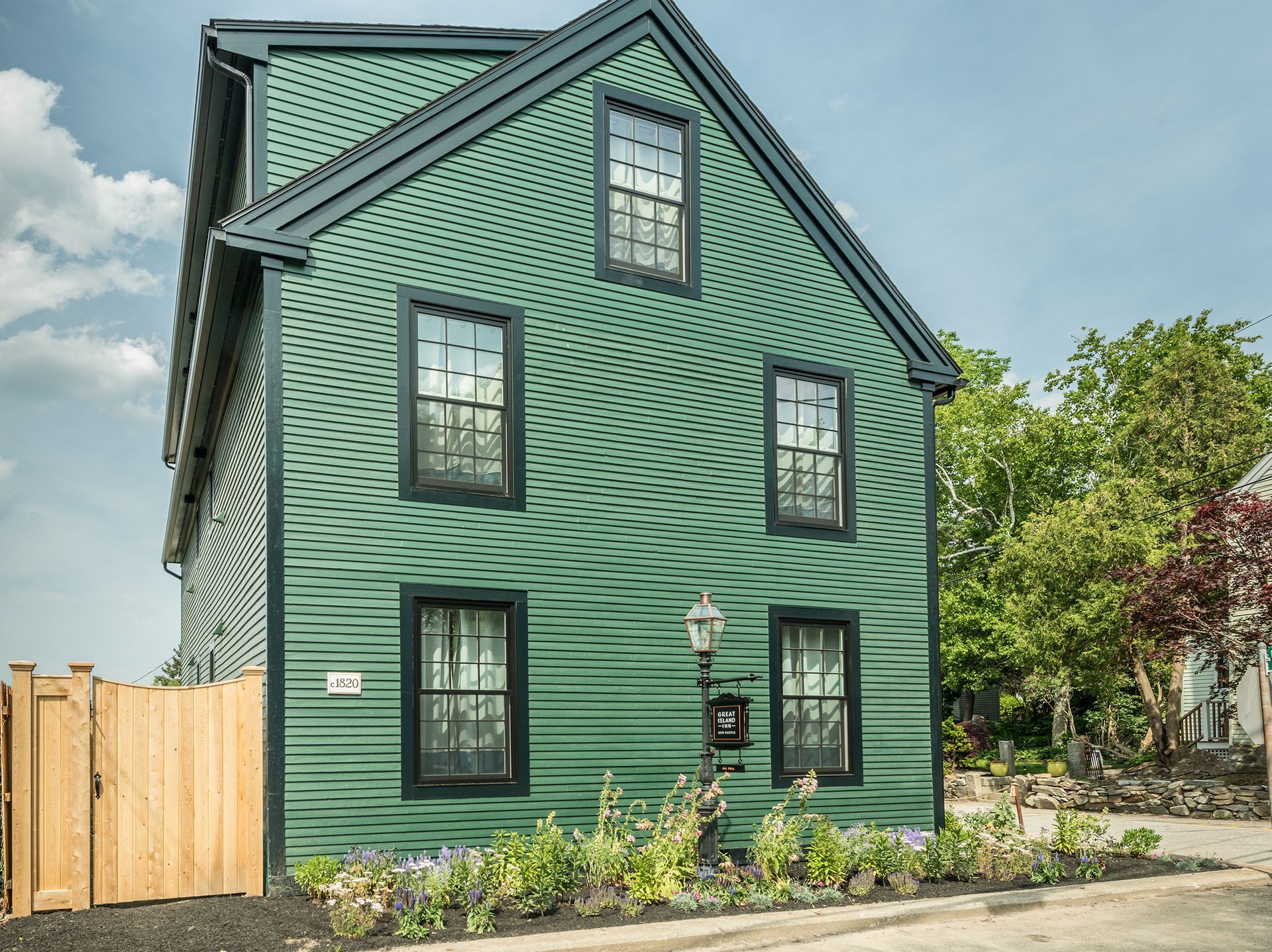 Green two-story house with dark trim, steep roof, and front garden beside a sidewalk
