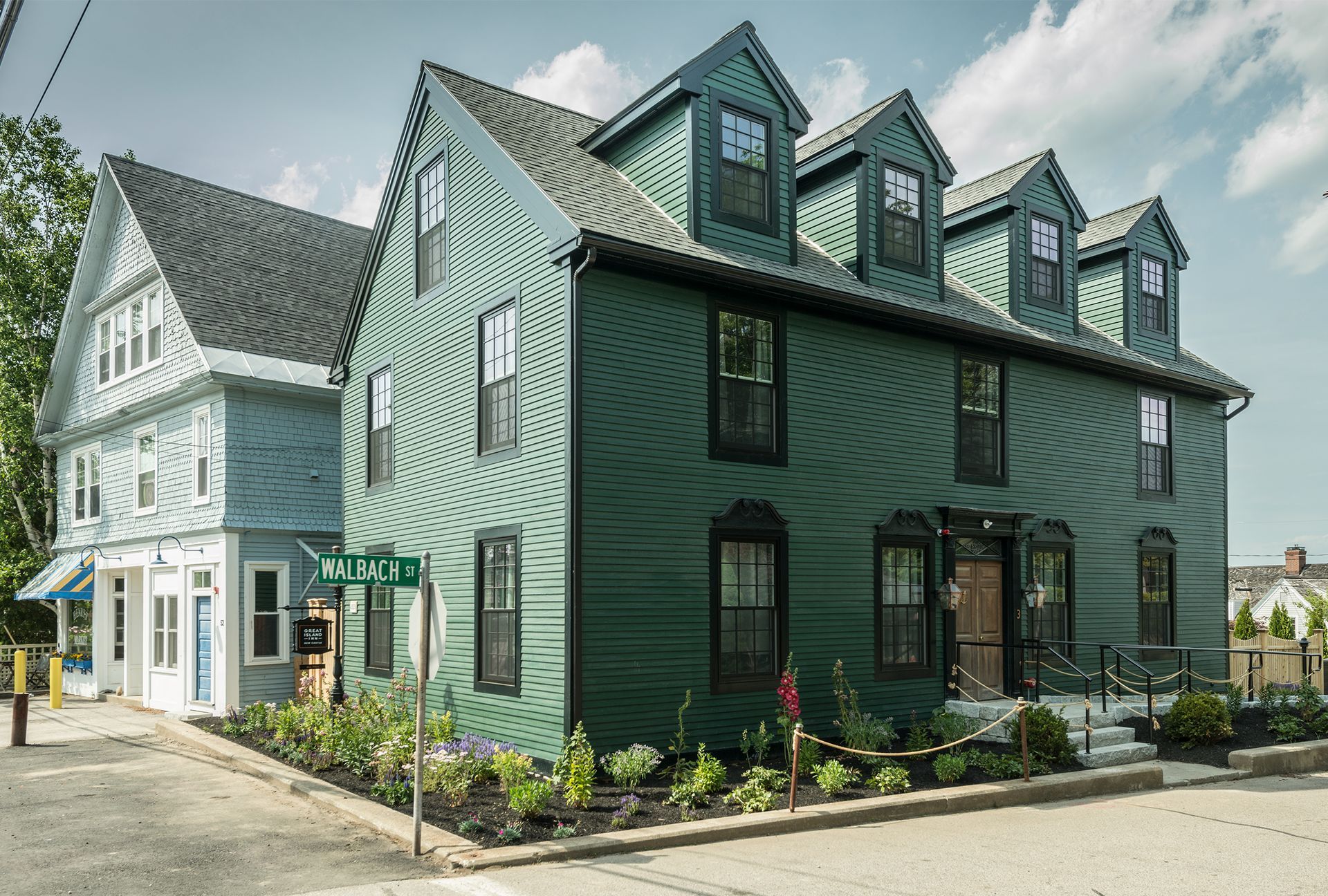 Two-story green house with dormer windows and porch beside a white building on a street corner