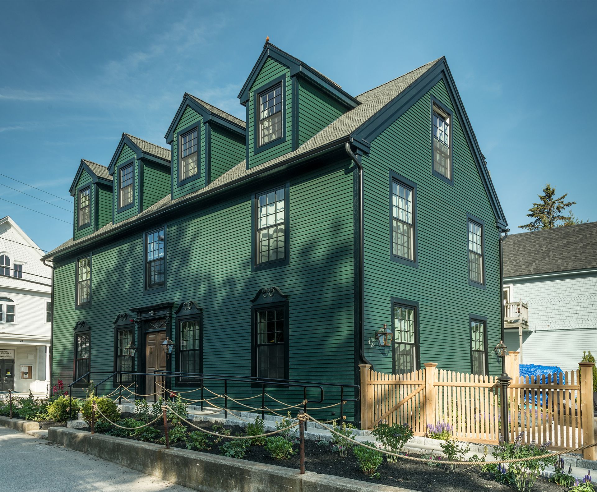 Green three-story house with dormer windows and a small fenced garden on a sunny street corner