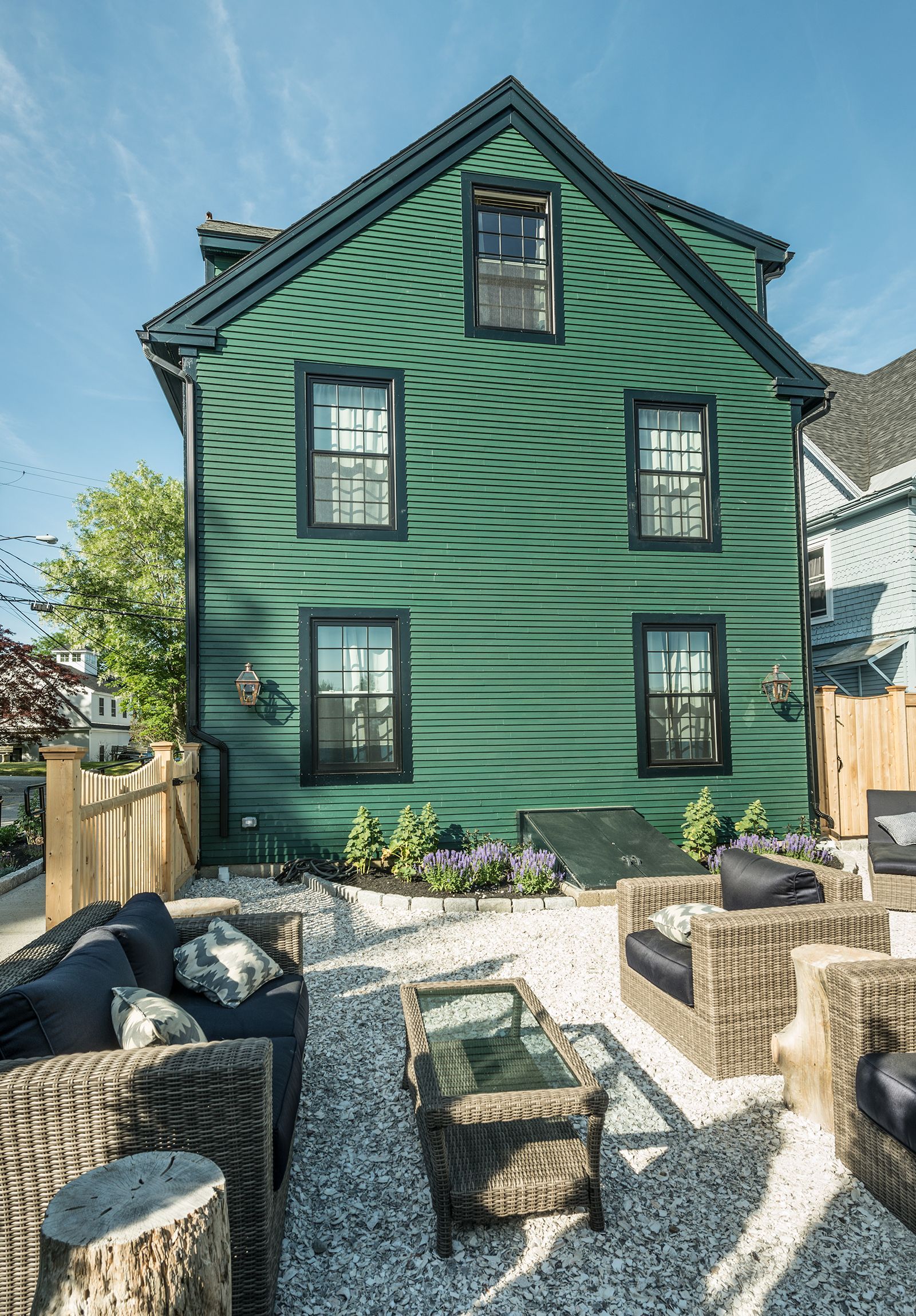 Green clapboard house with black trim and a gravel patio with wicker seating in front