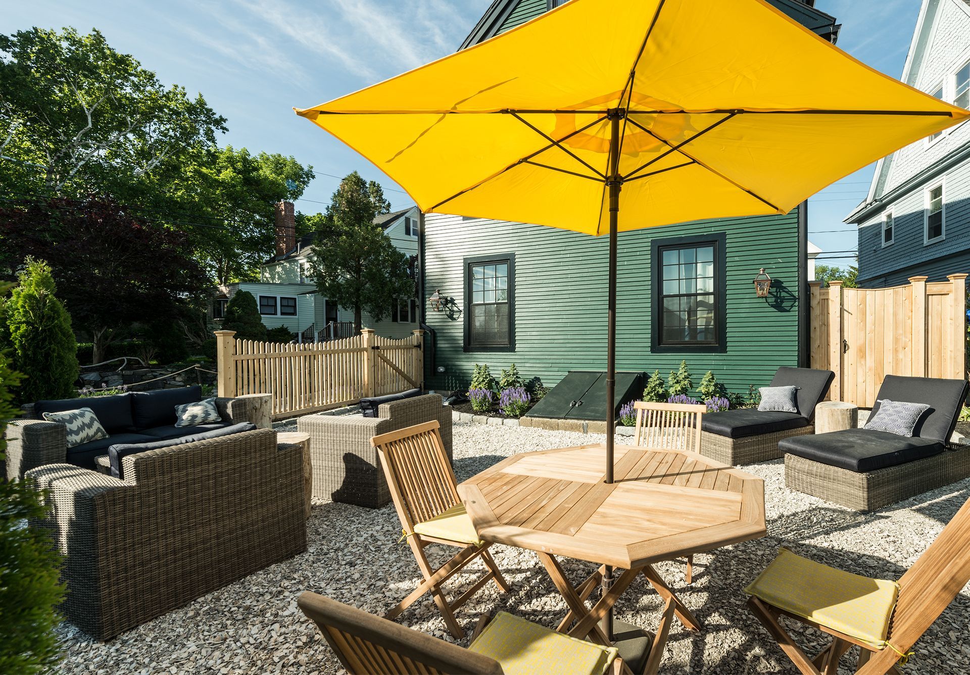 Sunny backyard patio with wicker seating, a wooden table, and a large yellow umbrella in front of a small shed