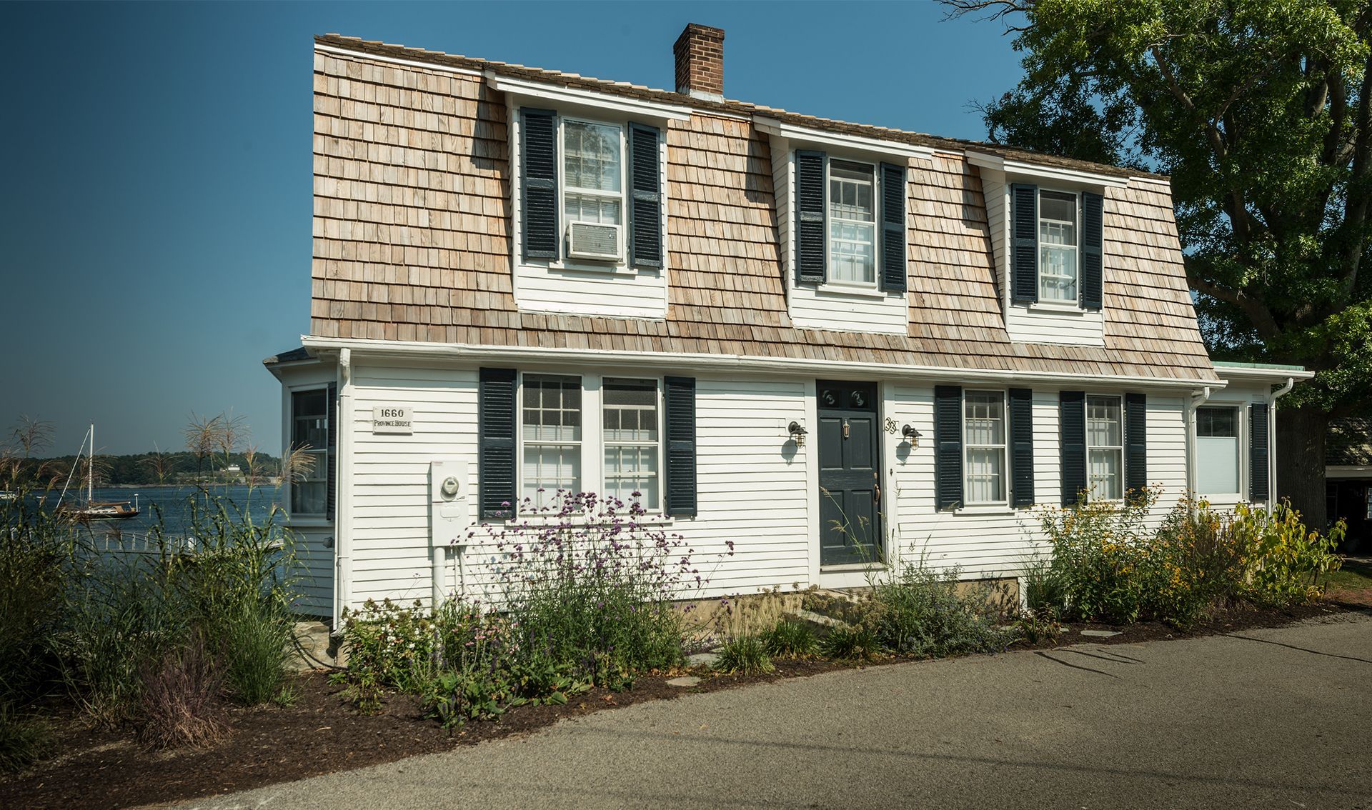 White waterfront cottage with dormer windows and a shingled roof beside a driveway