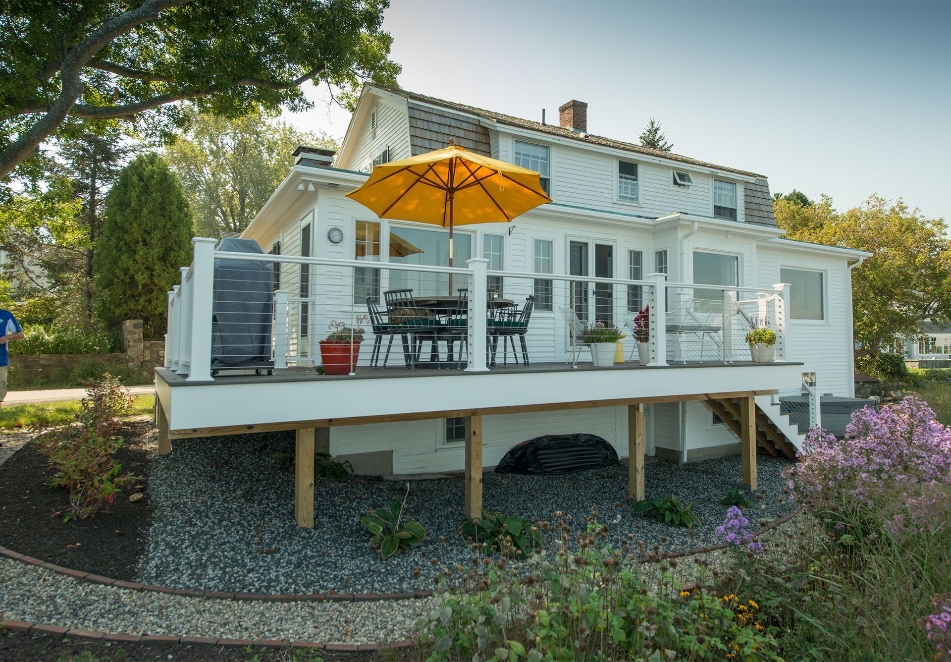 White house with a raised deck, orange umbrella, and garden landscaping surrounded by trees.