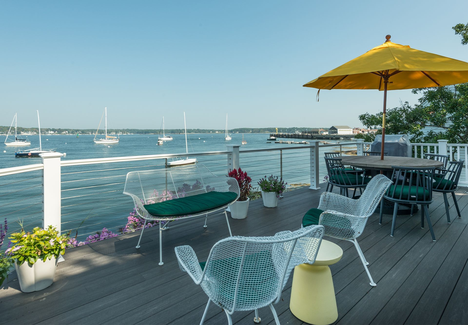 Waterfront deck with white chairs, yellow umbrella, and potted flowers overlooking boats on the water