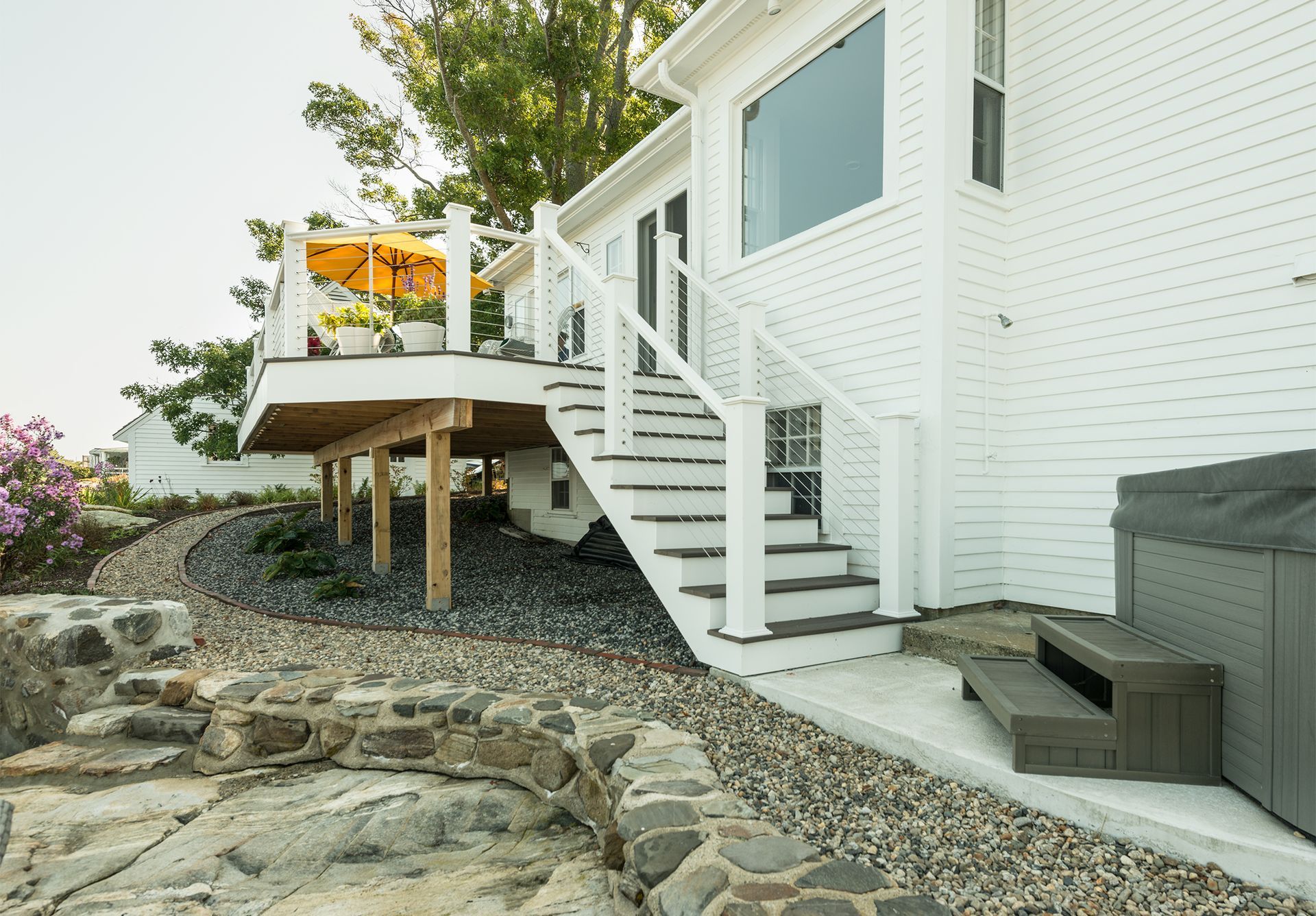 White house exterior with wooden deck stairs, white railing, and stone landscaping beside a rocky shore