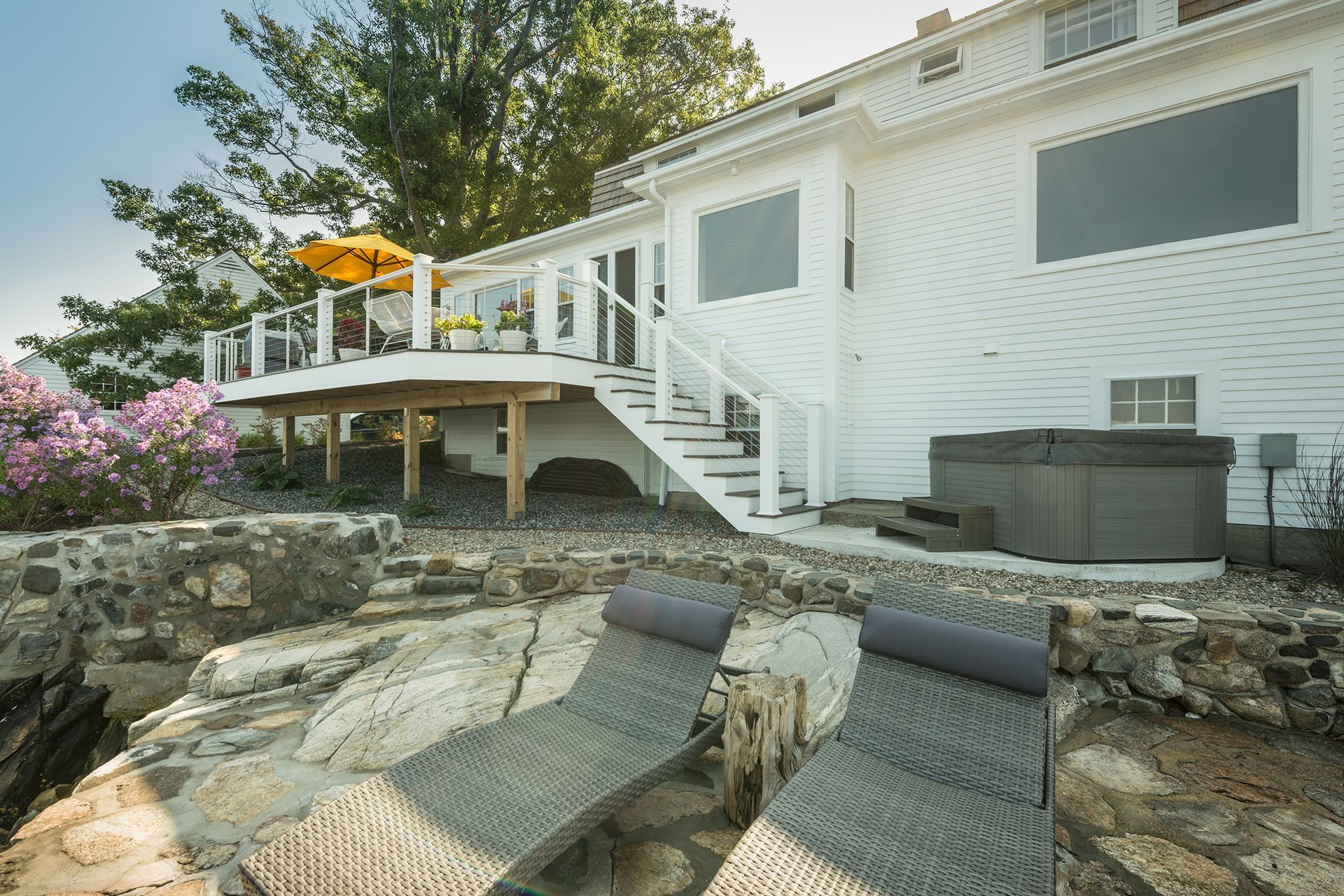 Backyard deck with white house, stairs, patio furniture, and rock landscaping overlooking water.