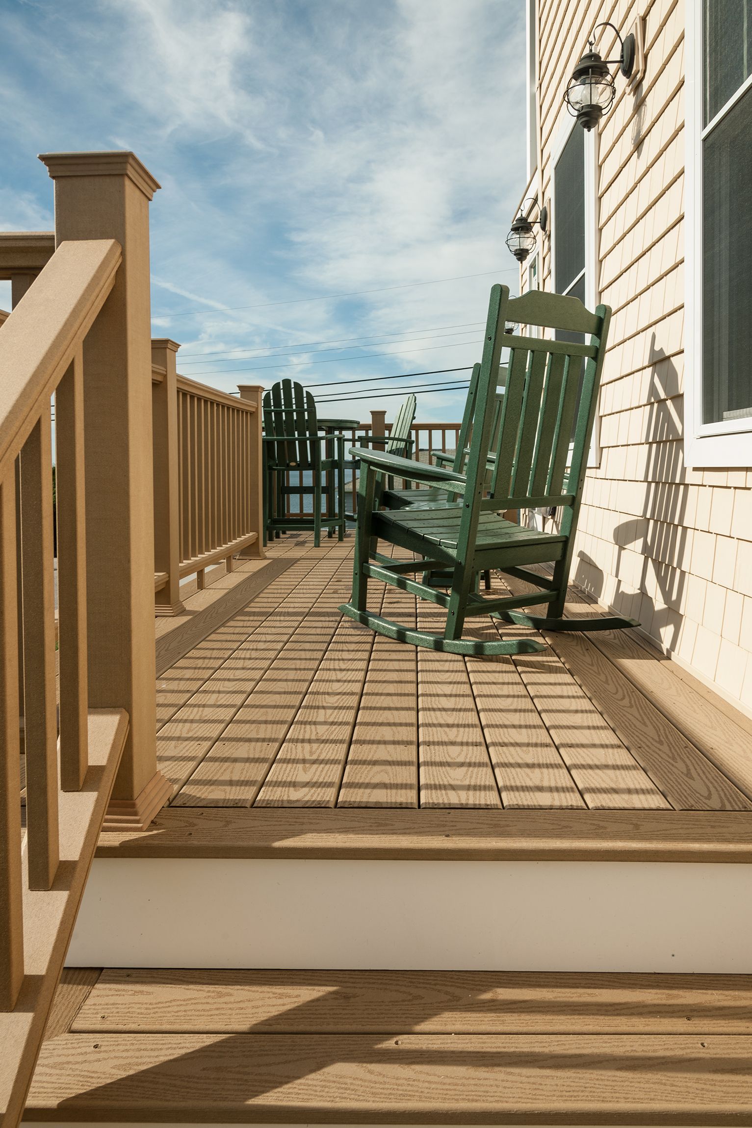 Wooden deck with green rocking chair beside a white house, overlooking a fence and ocean view.