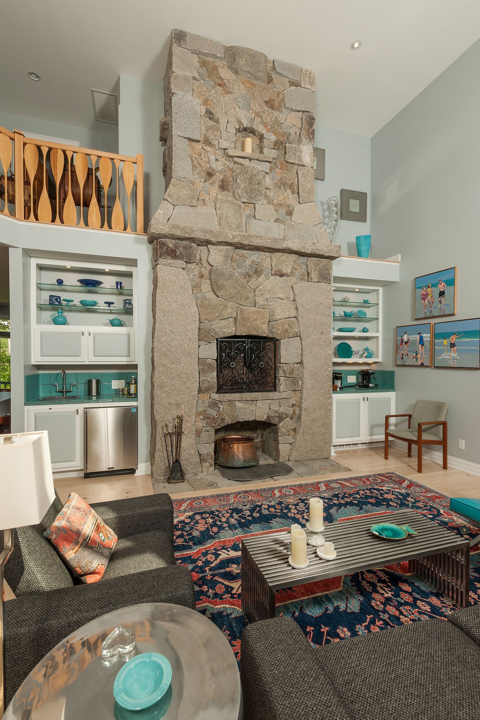 Cozy living room with a tall stone fireplace, colorful rug, shelves, and a glass coffee table with chess pieces.