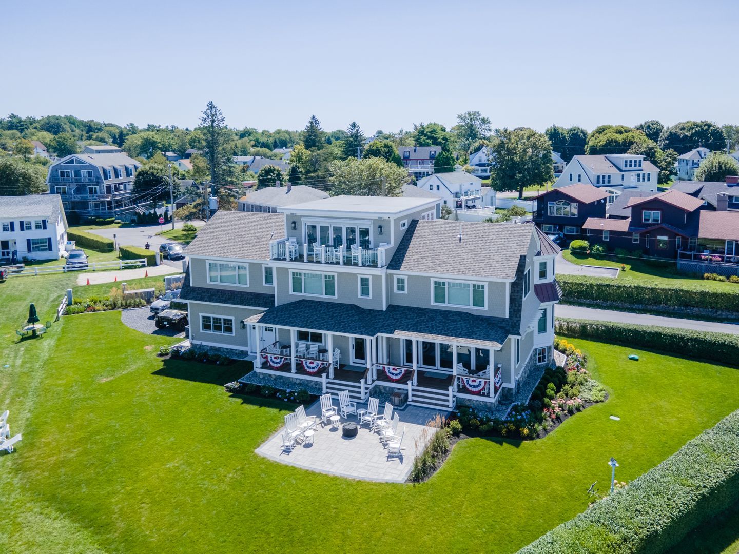 Large gray house with a wraparound porch and patio, set on a green lawn in a suburban neighborhood.