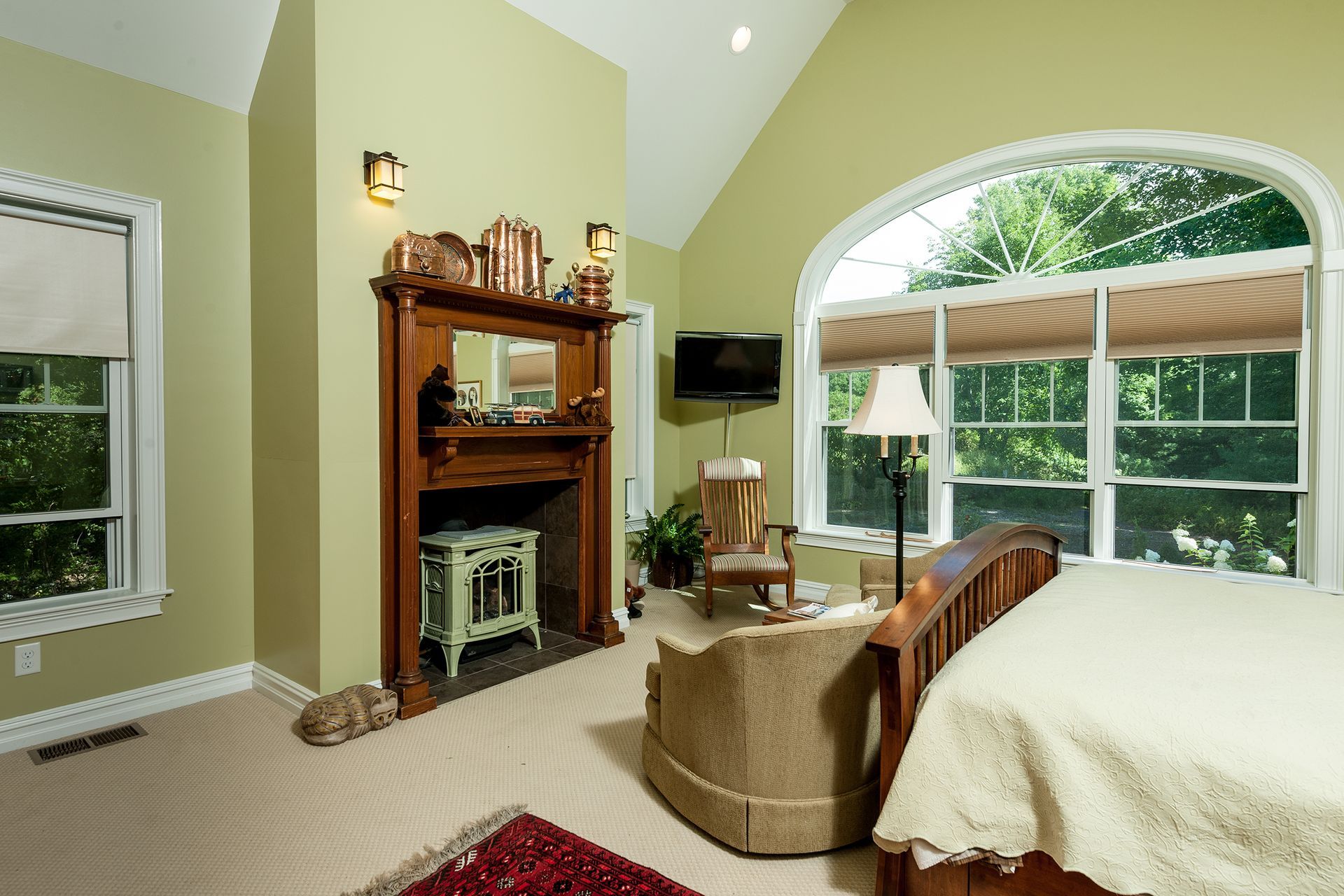 Bright bedroom with vaulted ceiling, fireplace, arched window, and bed beside a wicker chair.