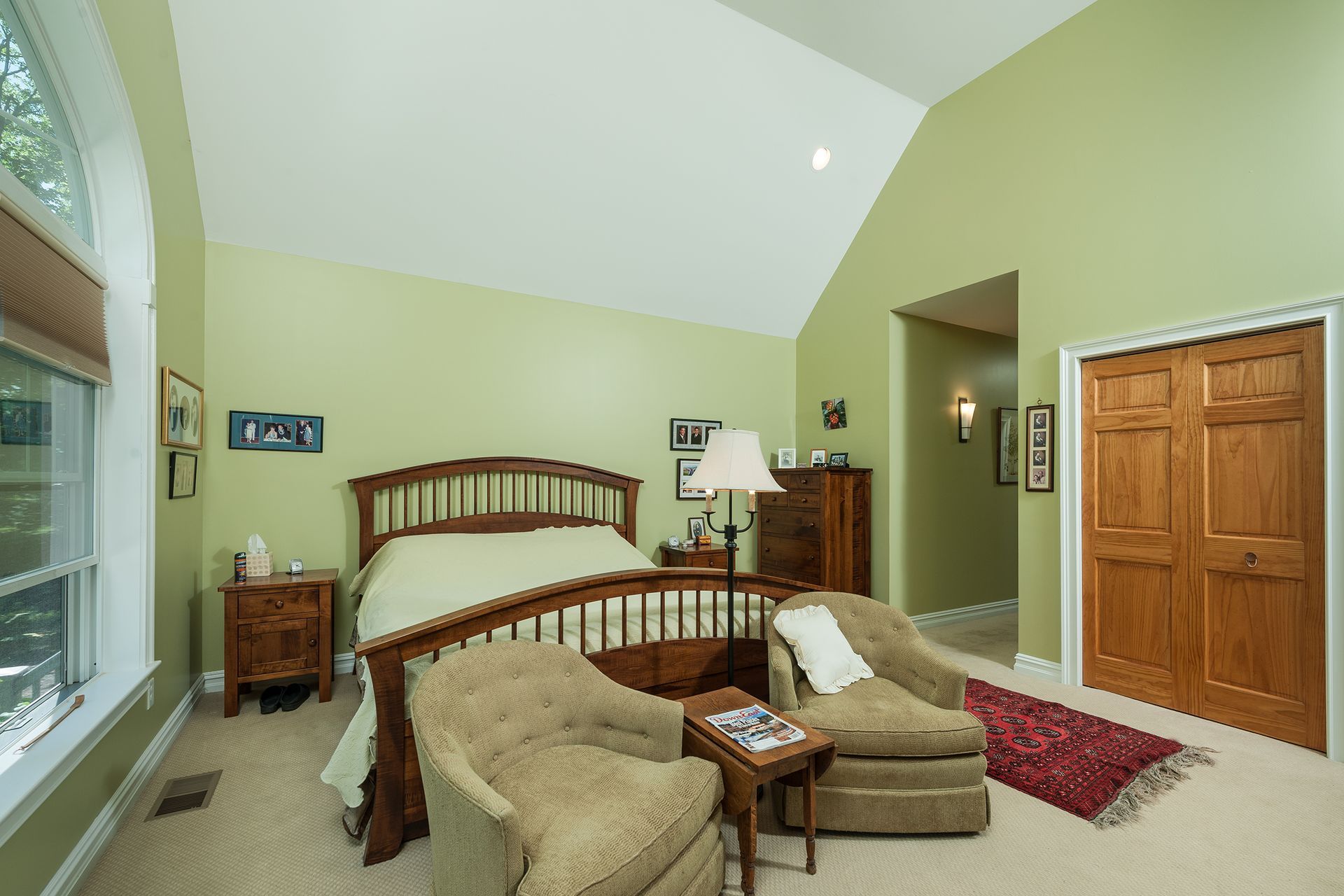 Bedroom with arched window, wood bed, beige chairs, red rug, and light green walls