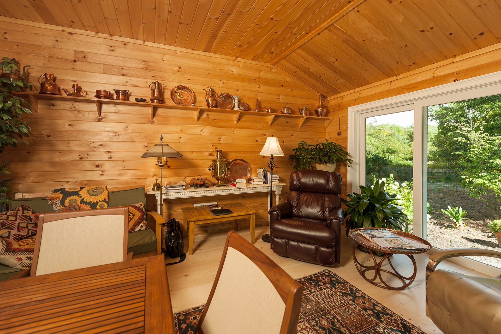 Wood-paneled sunroom with desk, leather chair, sofa, and open sliding doors to a green patio garden