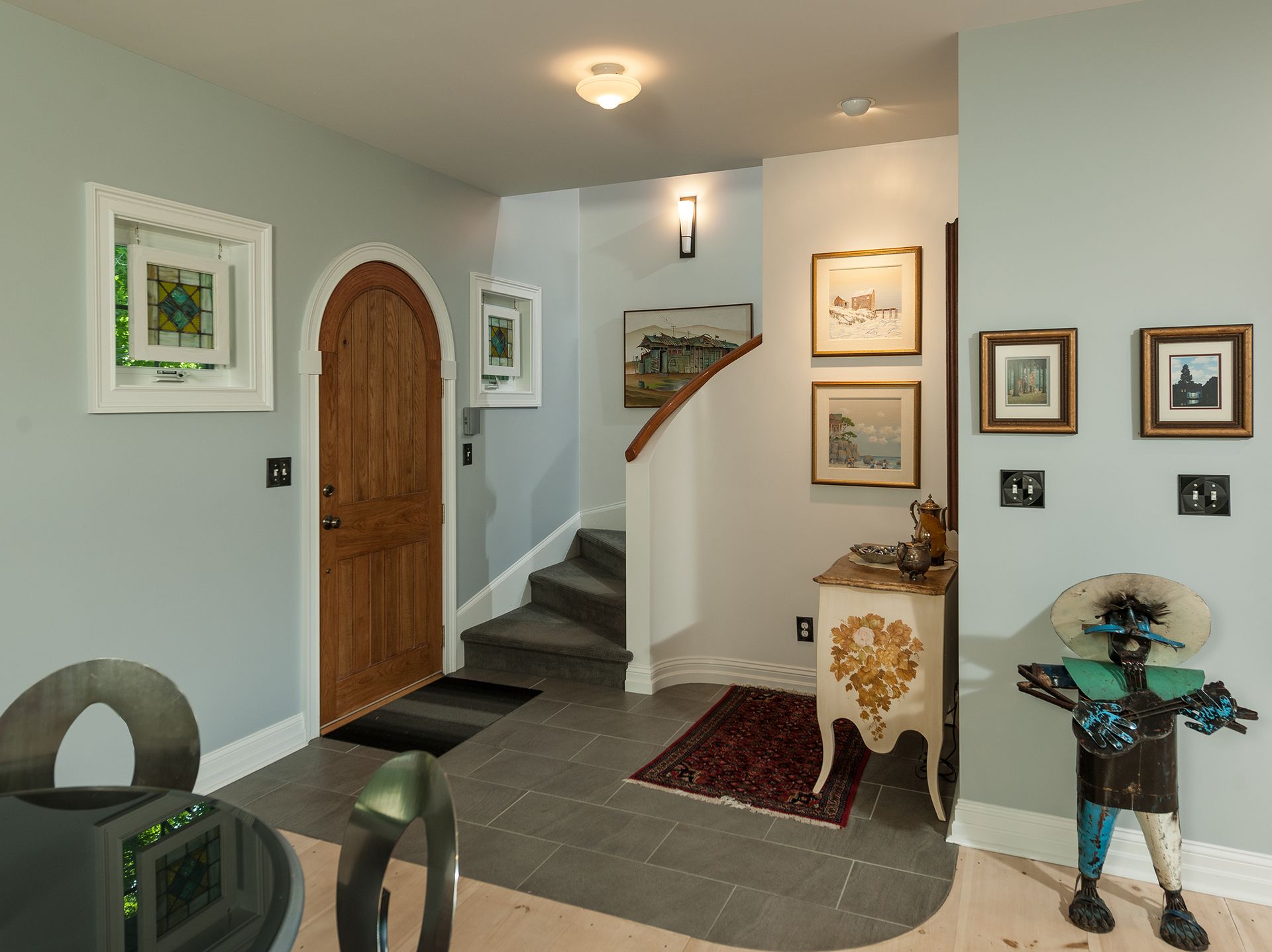 Hallway with arched wooden door, framed art, staircase, and decorative statue on a small rug