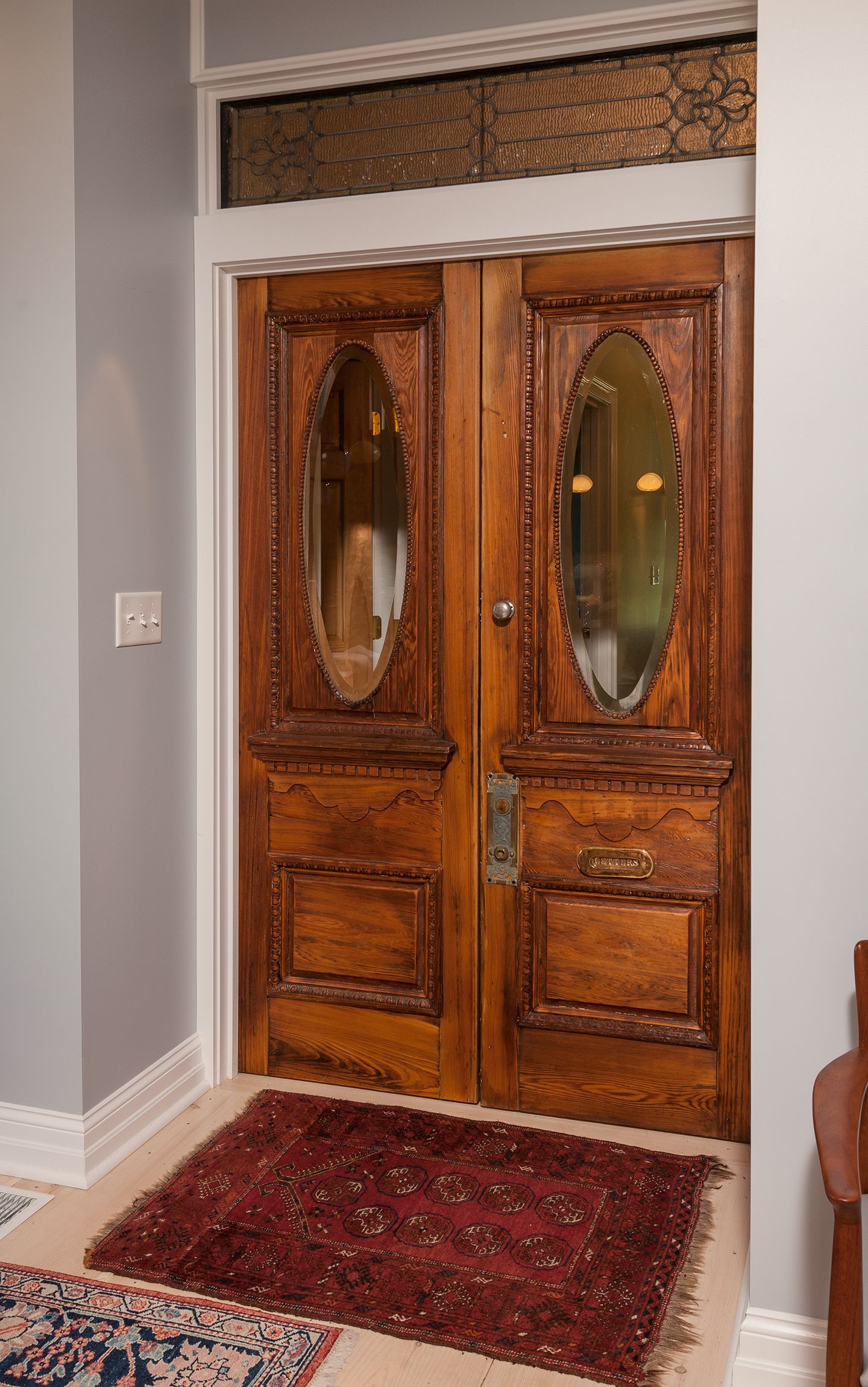 Wooden double front doors with oval glass panes, framed by gray walls and a red rug.