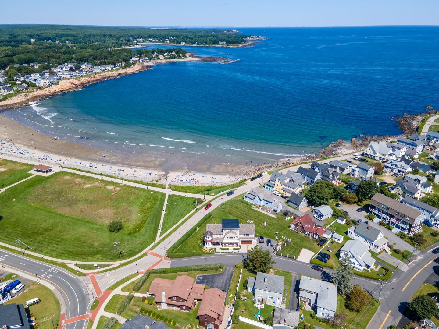 Aerial view of a coastal town with a curved beach, blue ocean, roads, and houses along the shoreline