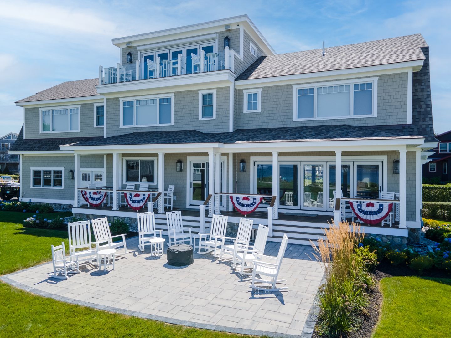 Large gray beach house with white trim and patriotic bunting, featuring a patio with white chairs and tables