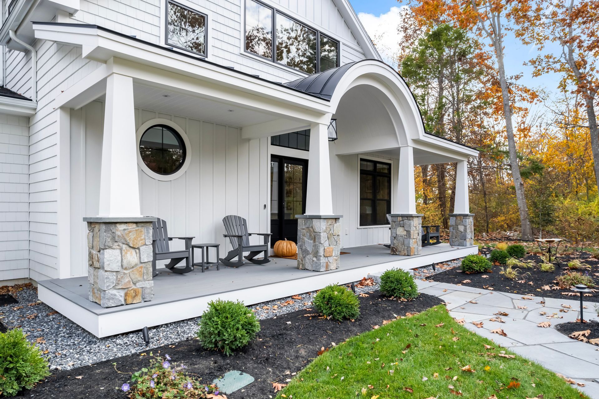 White house front porch with stone columns, black chairs, and autumn landscaping