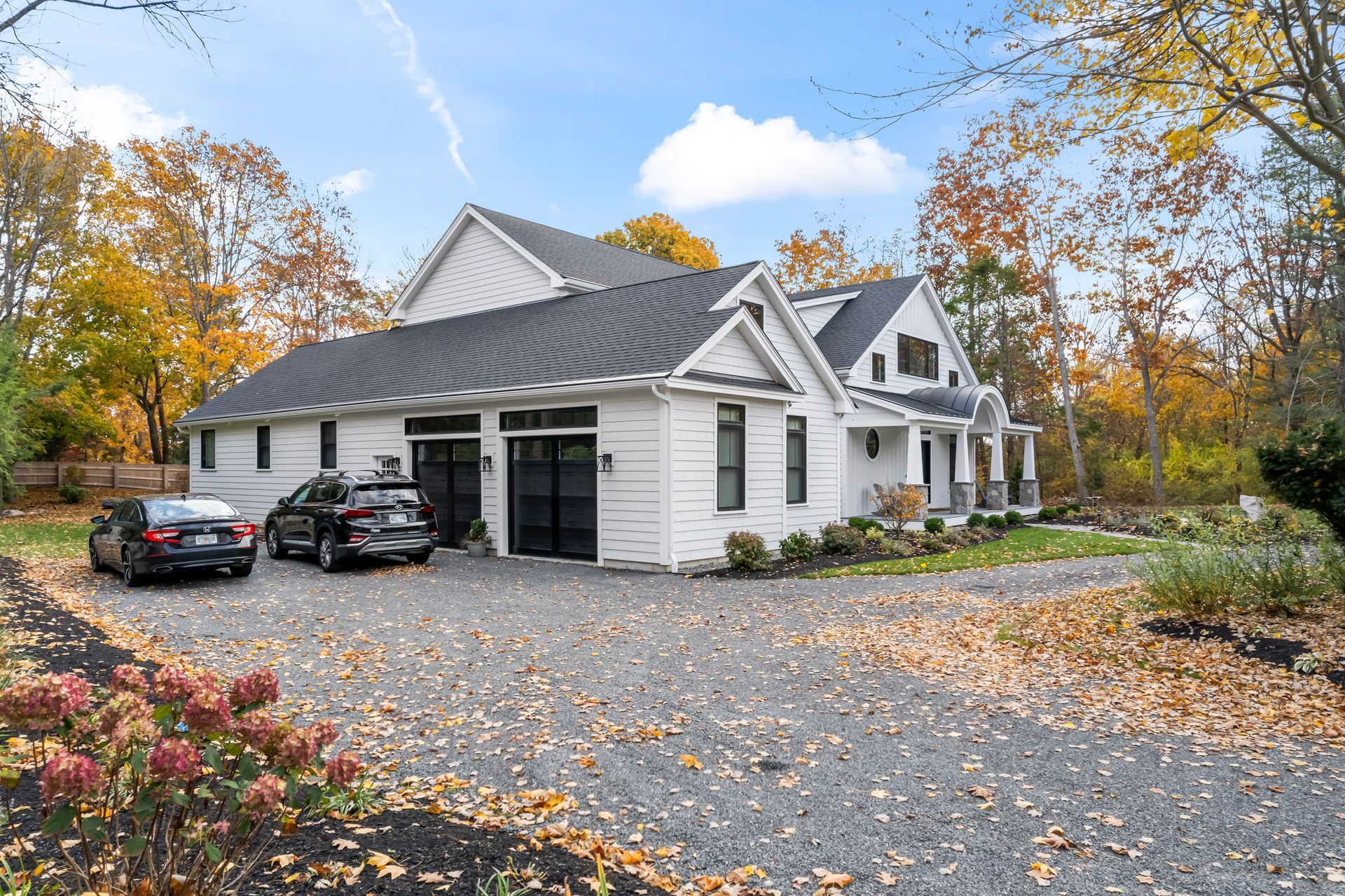 White house with gray roof and black cars on a gravel driveway, surrounded by autumn trees and fallen leaves