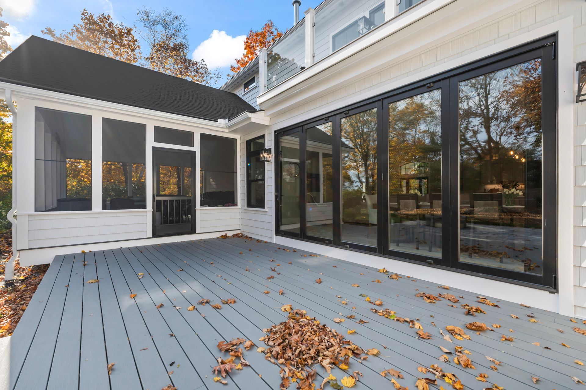 Modern house deck with gray boards, black-framed glass doors, and autumn leaves scattered outside