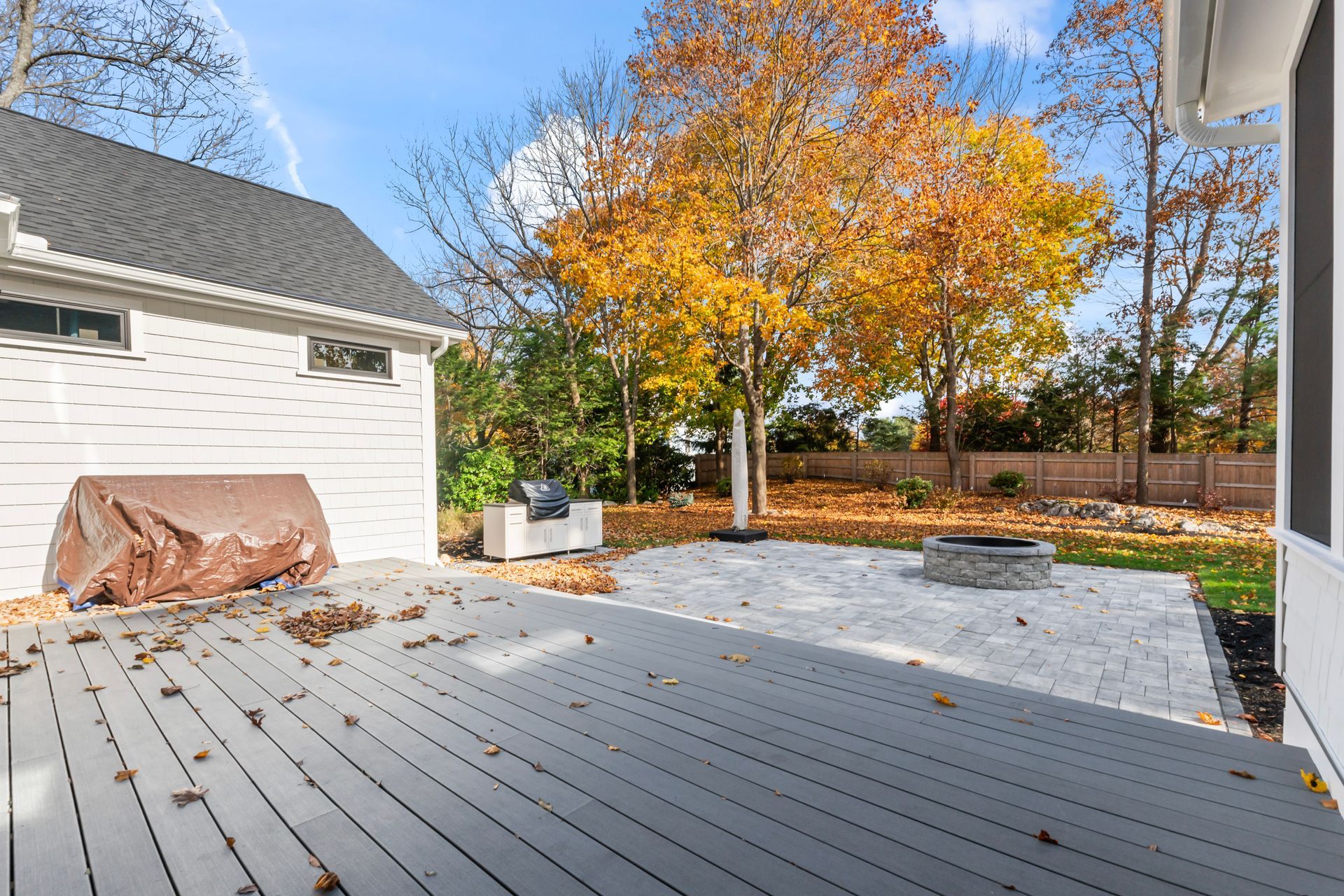 Backyard deck with white house siding, autumn trees, and a covered grill under a clear sky.