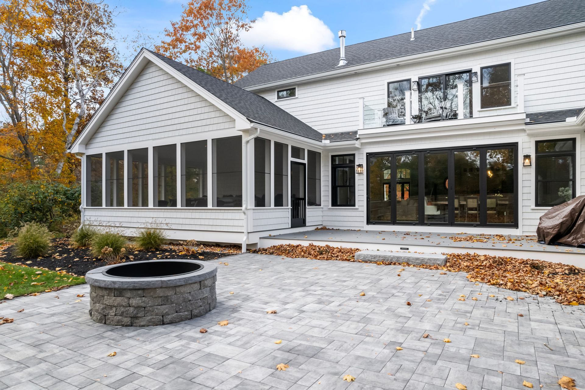 Backyard patio with white house, screened porch, paver terrace, and stone fire pit in autumn.