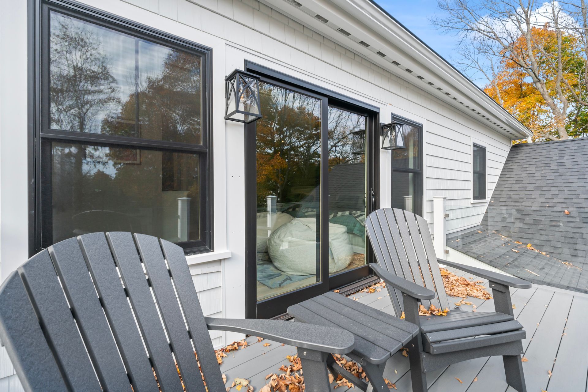 Two gray Adirondack chairs on a deck beside a white house with large windows and a glass door