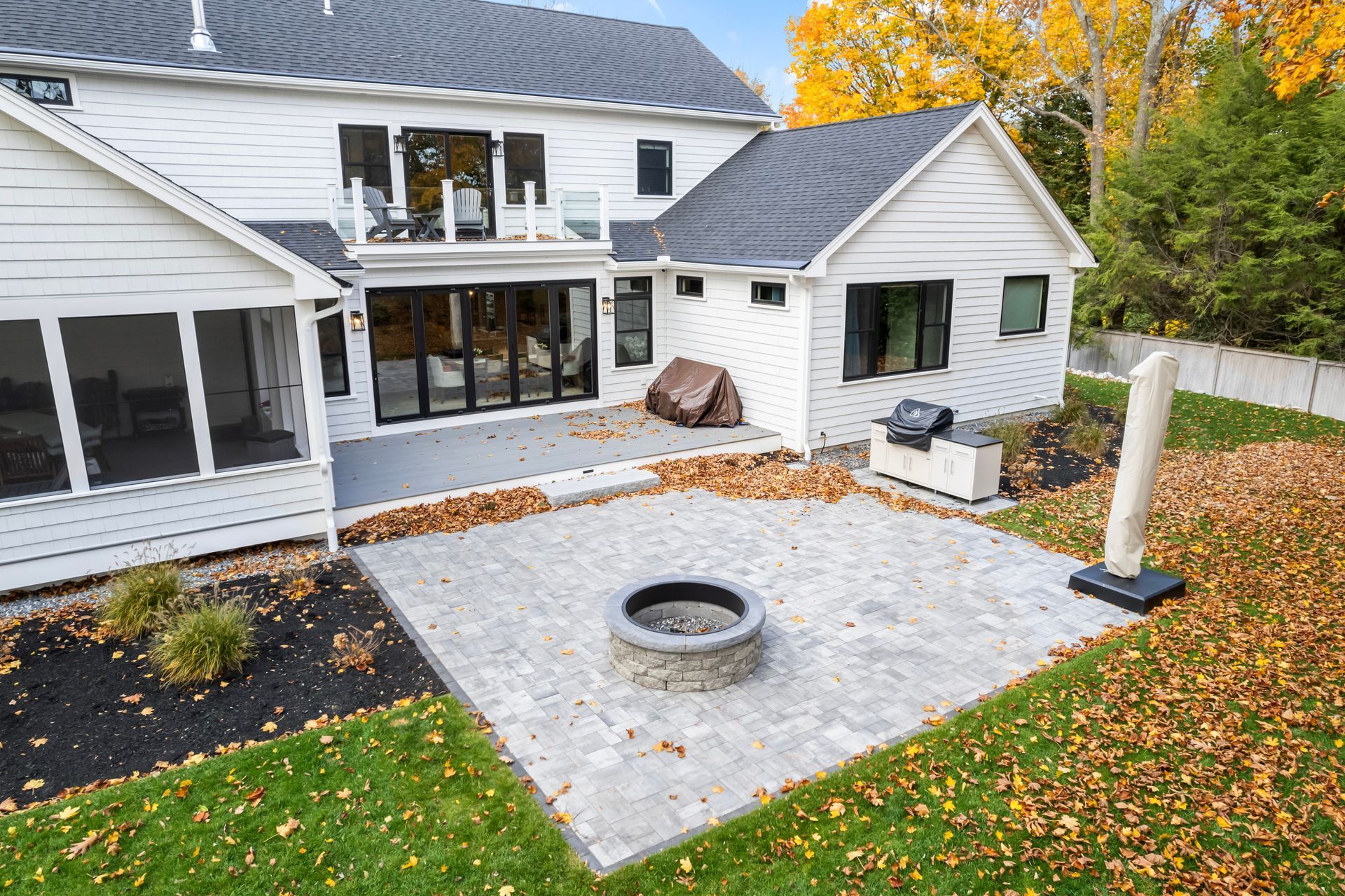 Modern white house with a stone patio and circular fire pit, surrounded by autumn leaves and landscaped yard