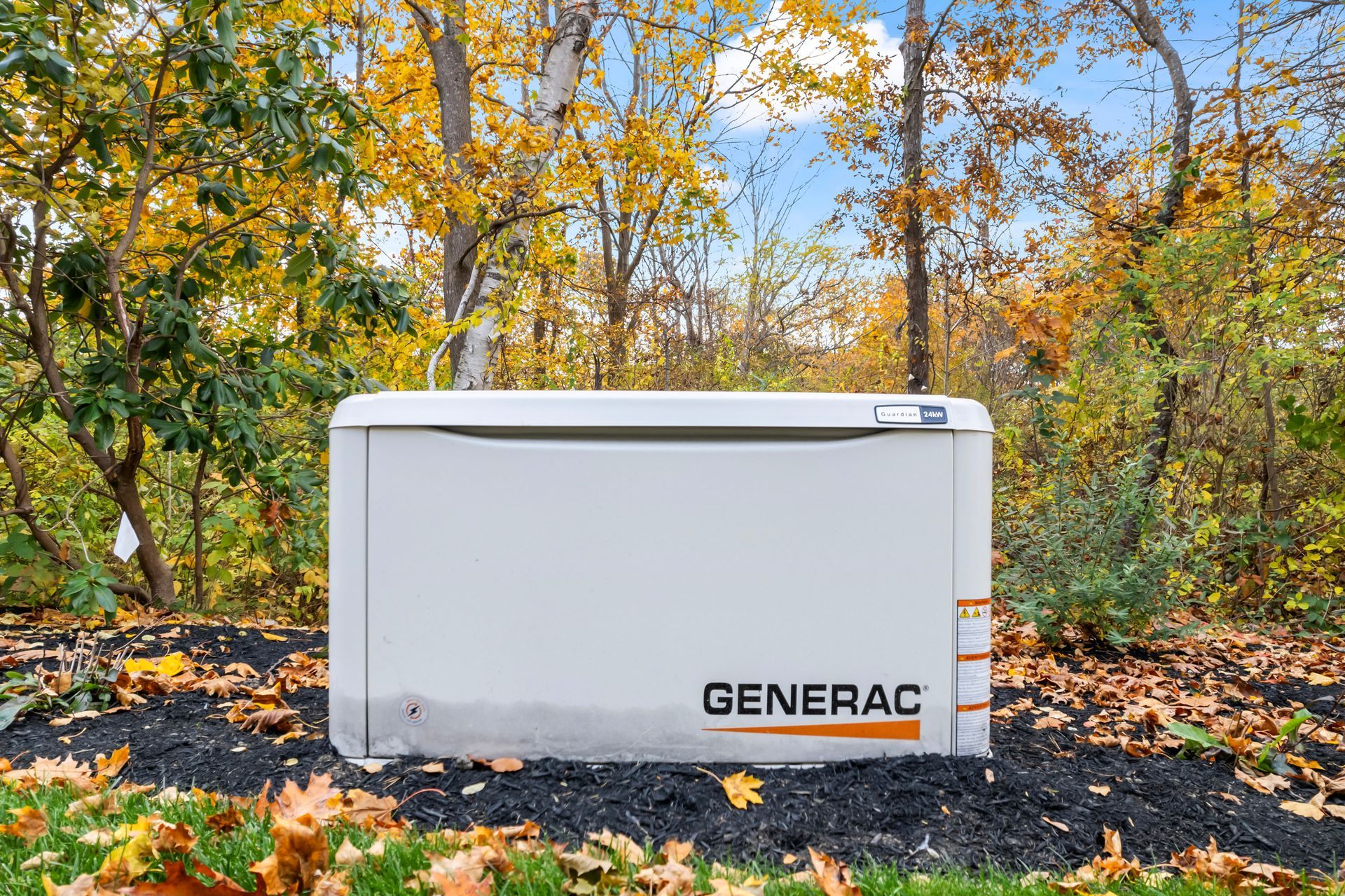 White Generac generator outdoors on a gravel pad, surrounded by autumn trees.