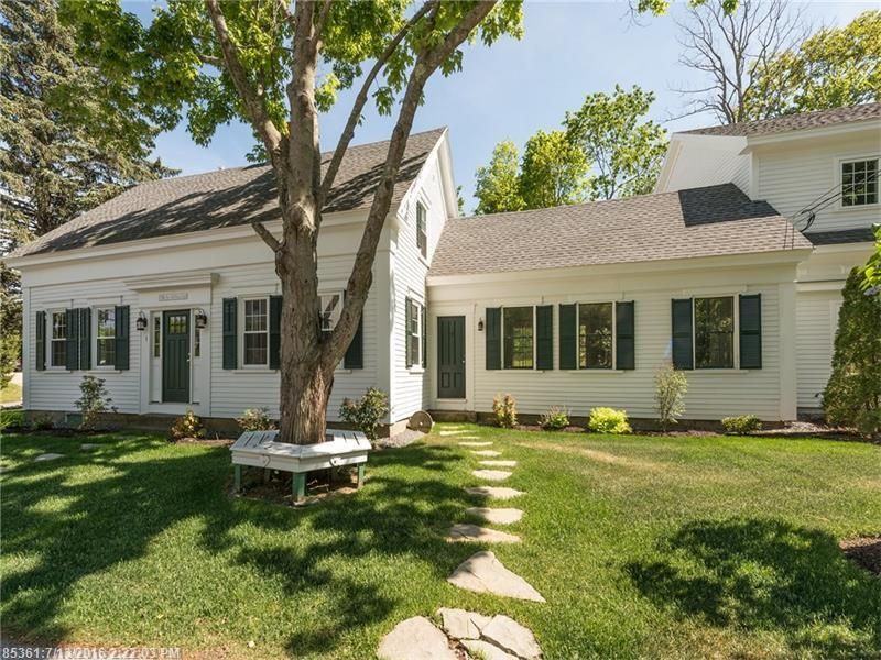 White house with green shutters and a front yard, shaded by large trees, with a stone path leading to the door