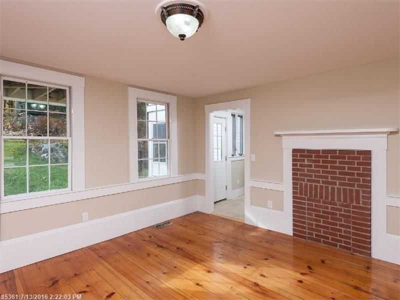 Empty room with hardwood floors, white trim, windows, and a brick fireplace.