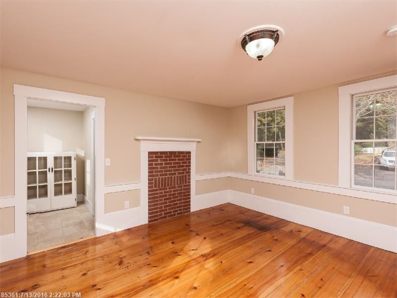 Empty room with hardwood floors, white trim, brick fireplace, and two windows.