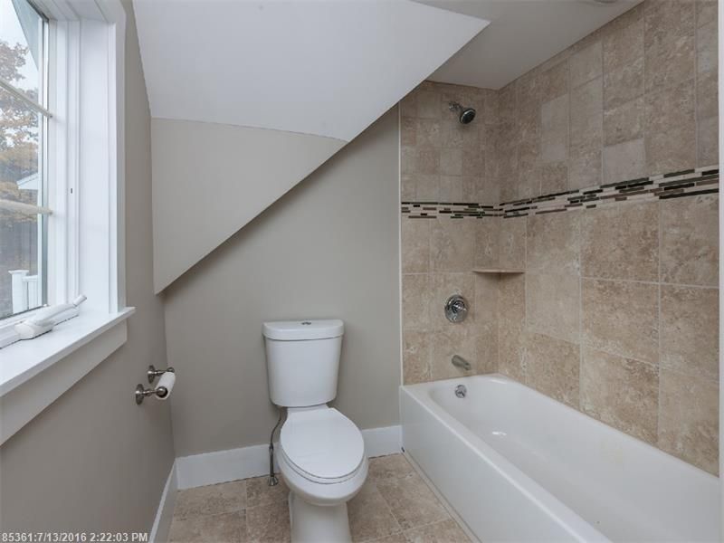 Small bathroom with sloped ceiling, toilet by window, and tiled bathtub/shower.