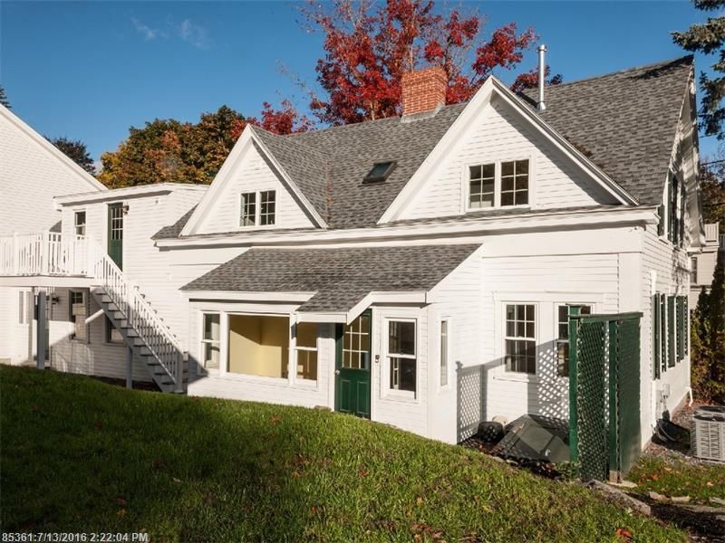White two-story house with gray roof and dormers, front porch, and green lawn in fall sunlight