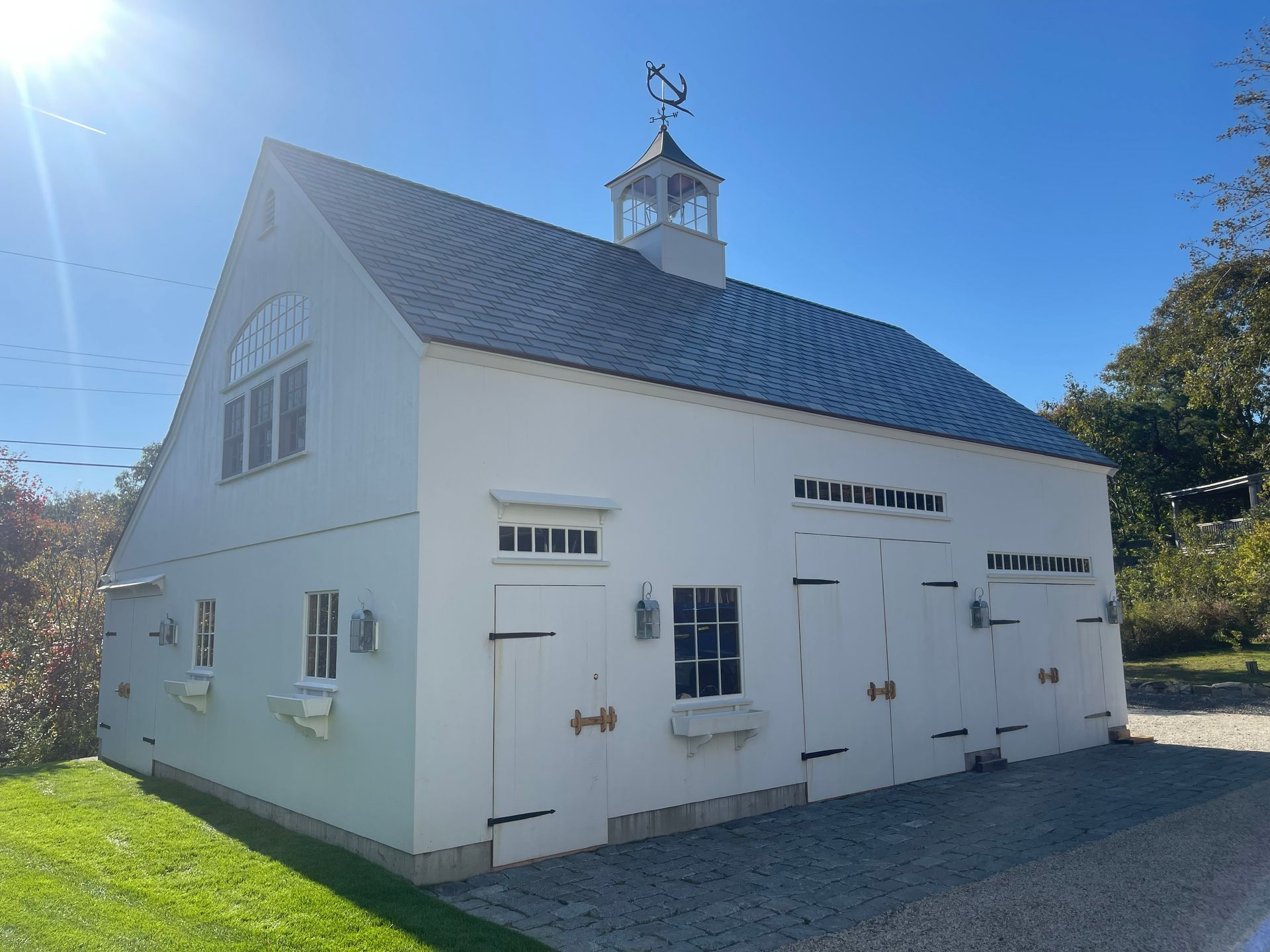 White barn-style building with a steep gray roof, cupola, and white doors beside a paved drive under a blue sky