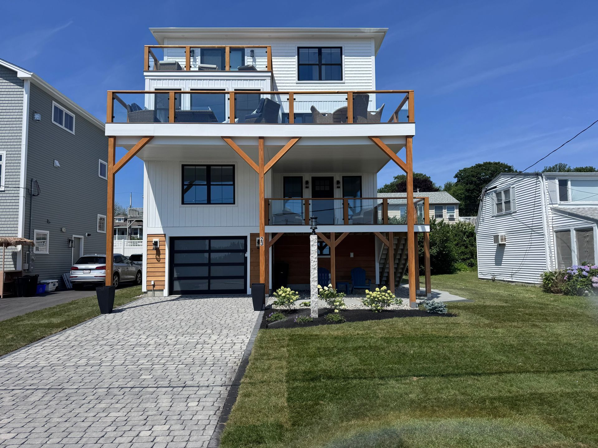 Modern white beachfront house with wooden decks, large windows, and a stone driveway under a clear blue sky
