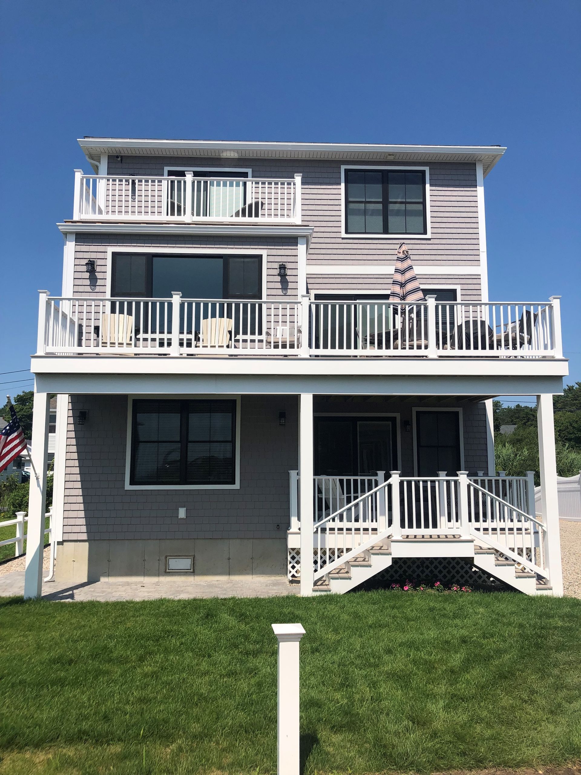 Three-story coastal house with white balconies, dark siding, and a front lawn under a clear blue sky