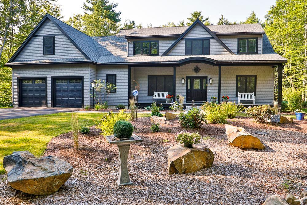 Gray house with black roof and front porch, set behind a gravel yard with large decorative boulders.