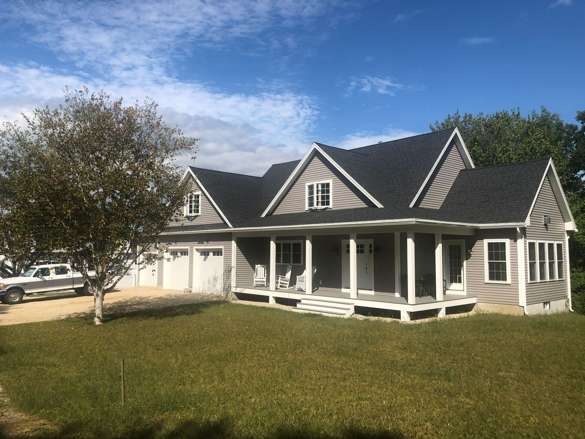 Single-story gray house with white porch and garage, front lawn, and a large tree under a blue sky