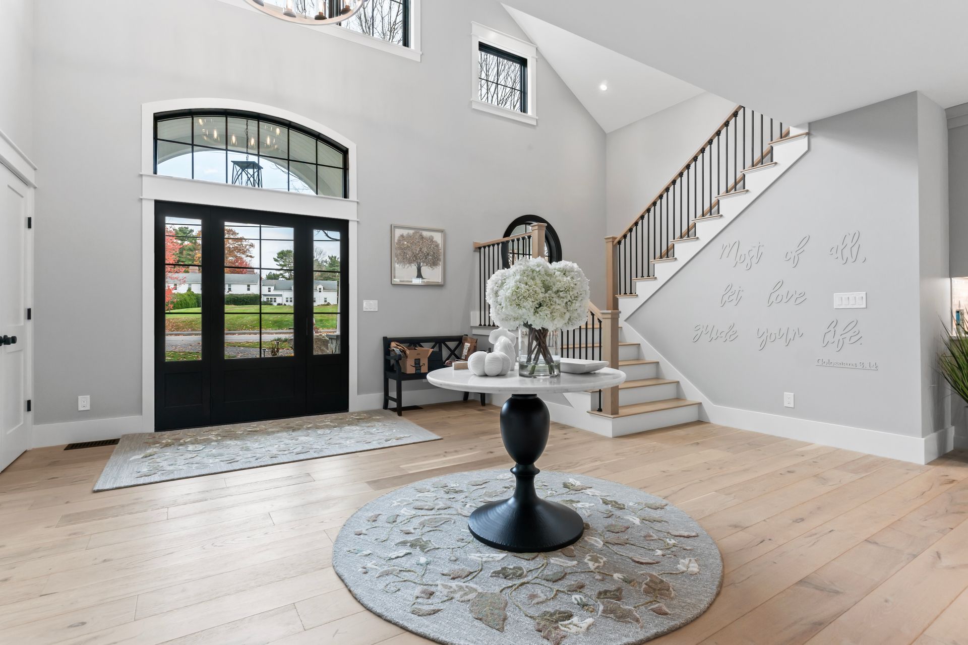 Bright foyer with black front door, staircase, light wood floors, and round table with flowers