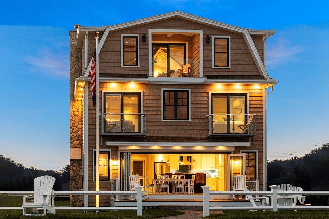 Two-story gray beach house lit warmly at dusk, with a white fence and ocean view.
