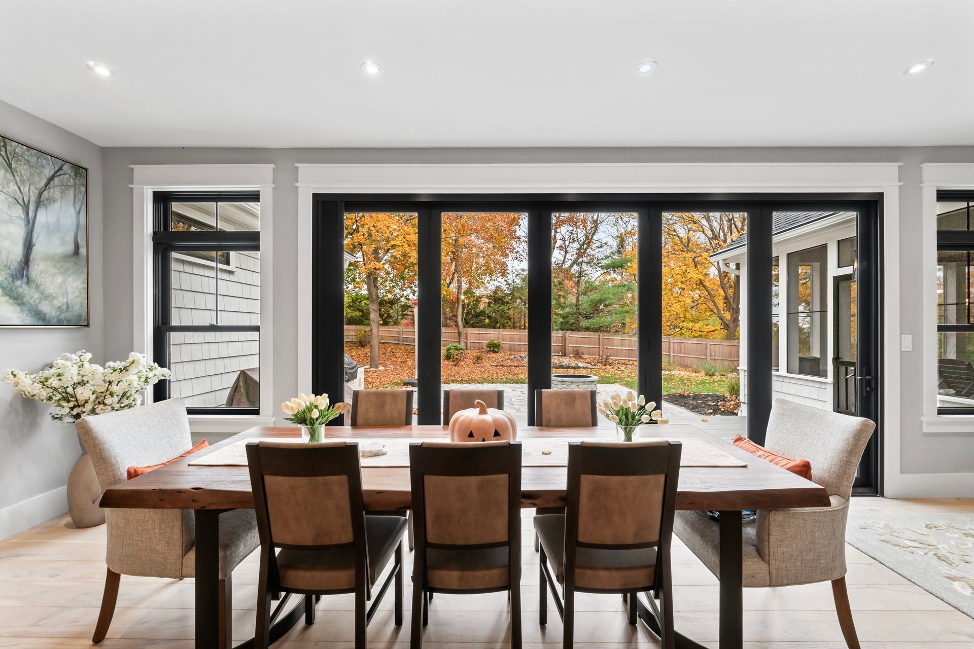 Modern dining room with a wooden table, chairs, and large windows overlooking autumn trees