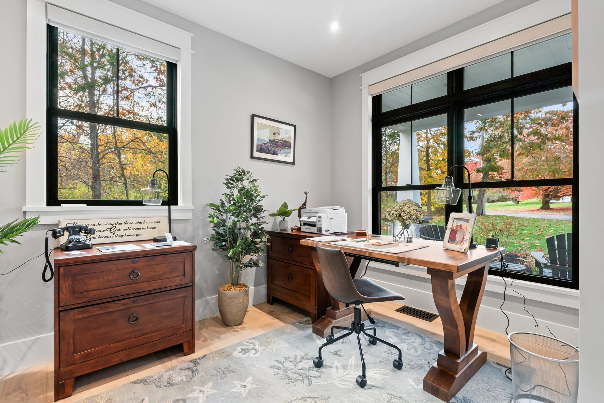 Bright home office with desk, chair, potted plant, and large windows overlooking autumn trees.
