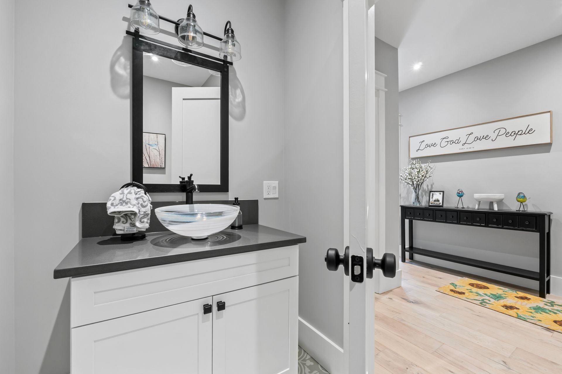 Small black-and-white powder room with vanity and mirror opening to a bright hallway.