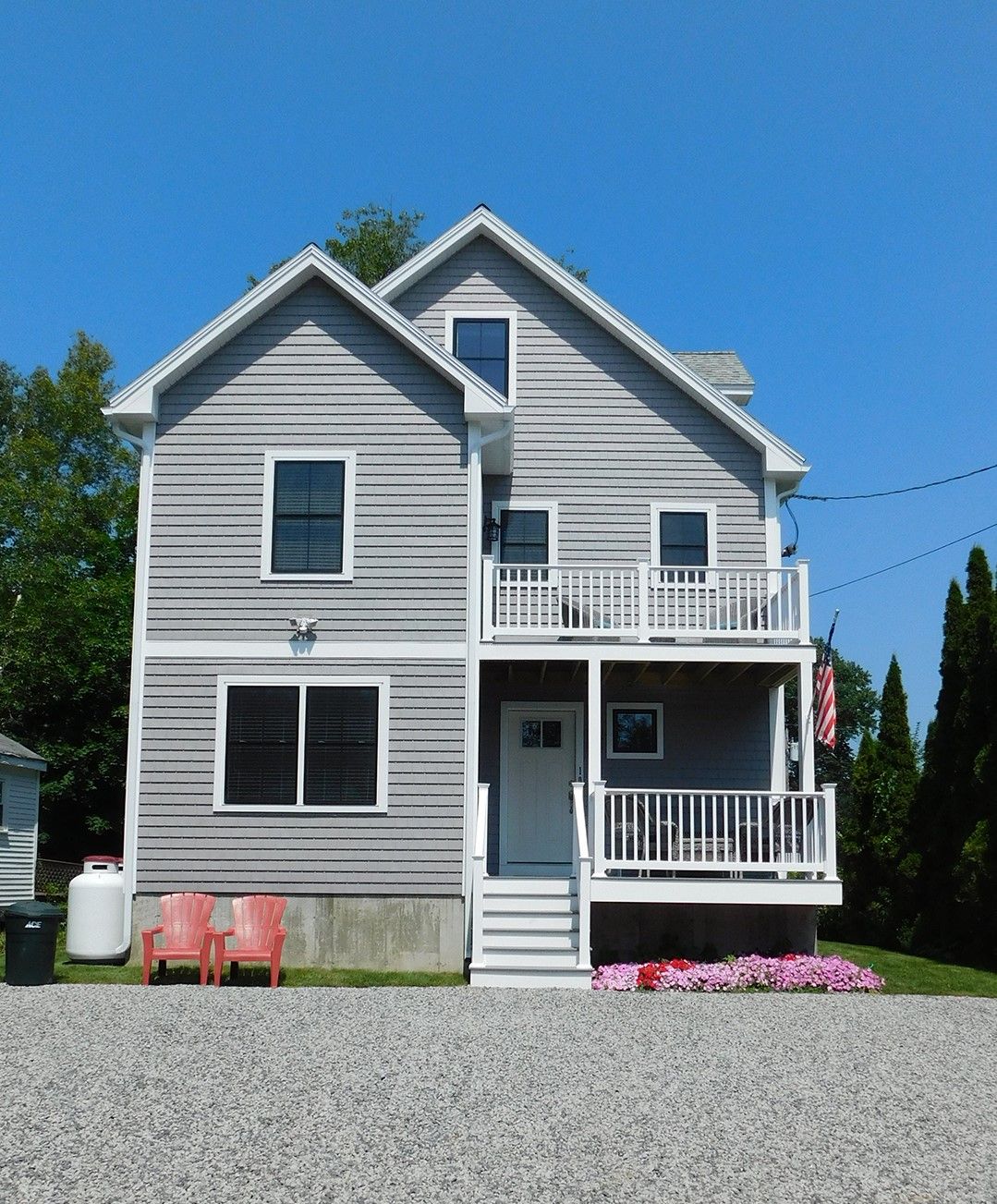 Gray two-story house with white trim, small porch, balcony, and gravel driveway under a blue sky