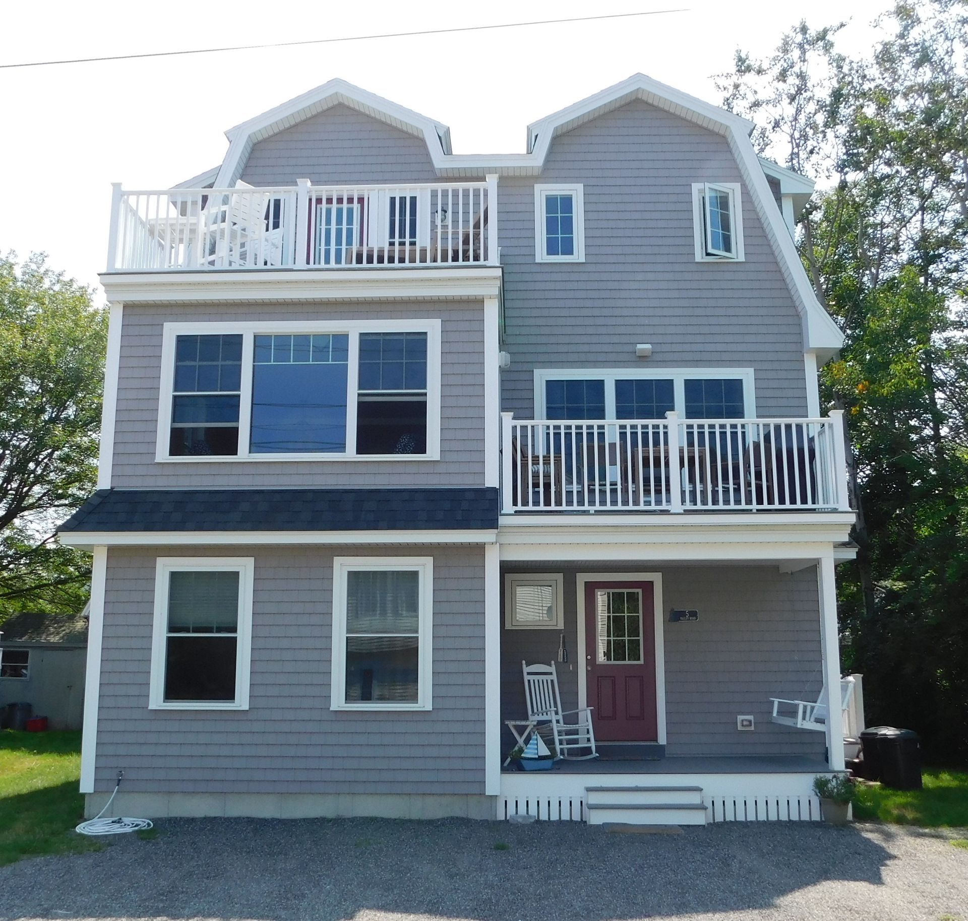 Gray two-story house with white trim, balconies, and a red front door on a gravel driveway