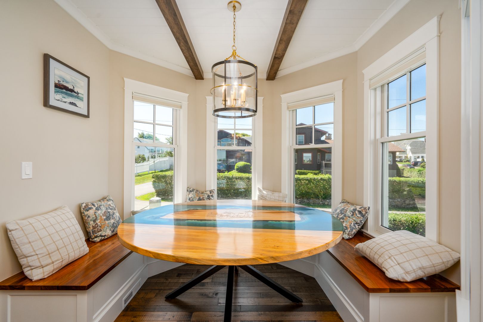 Bright breakfast nook with round wooden table, built-in bench seating, and large windows overlooking a yard